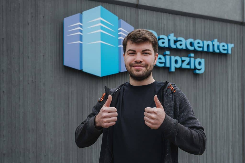 Azubi Edwin Schiewe steht vor seinem Ausbildungsbetrieb, dem Datacenter Leipzig 2, und zeigt beide Daumen nach oben 