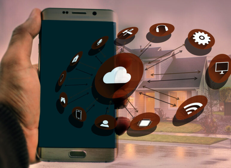 Hand hält Smartphone, überlagert mit Cloud- und Smart‑Home-Symbolen, Pfeile verbinden Geräteikonen vor einem Wohnhaus im Hintergrund