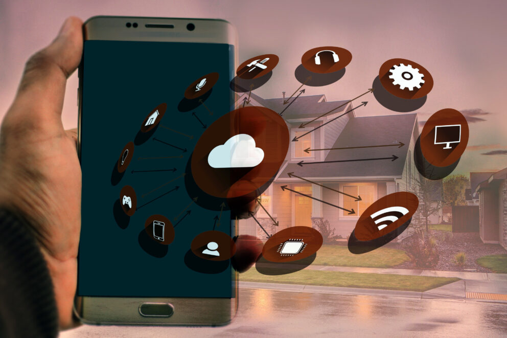 Hand hält Smartphone, überlagert mit Cloud- und Smart‑Home-Symbolen, Pfeile verbinden Geräteikonen vor einem Wohnhaus im Hintergrund