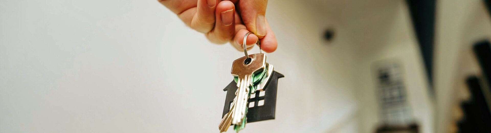 Hand hält Schlüsselbund mit schwarzem Haus-Anhänger, im Hintergrund unscharfe Treppe und Geländer.