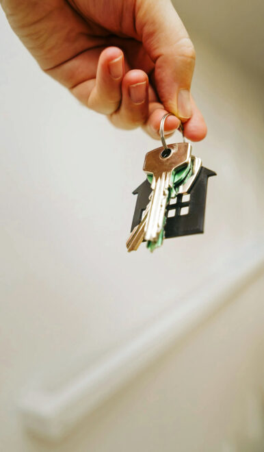 Hand hält Schlüsselbund mit schwarzem Haus-Anhänger, im Hintergrund unscharfe Treppe und Geländer.