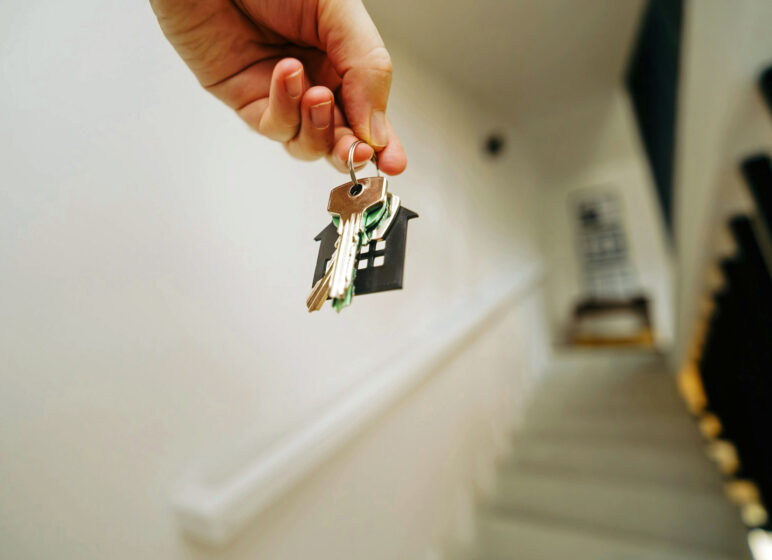 Hand hält Schlüsselbund mit schwarzem Haus-Anhänger, im Hintergrund unscharfe Treppe und Geländer.