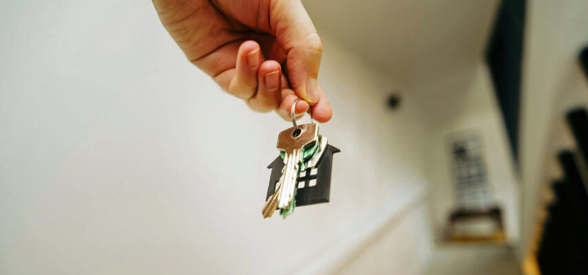 Hand hält Schlüsselbund mit schwarzem Haus-Anhänger, im Hintergrund unscharfe Treppe und Geländer.