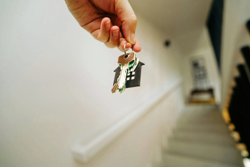 Hand hält Schlüsselbund mit schwarzem Haus-Anhänger, im Hintergrund unscharfe Treppe und Geländer.