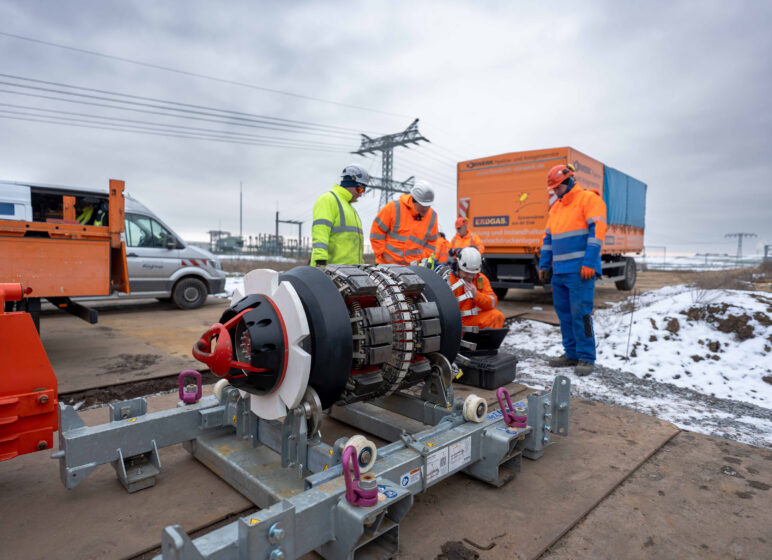 Arbeiter in Warnschutzkleidung prüfen ein großes Rohrbohrwerk auf einer verschneiten Baustelle vor orangefarbenem Firmen-Lkw und Stromleitungen im Hintergrund.