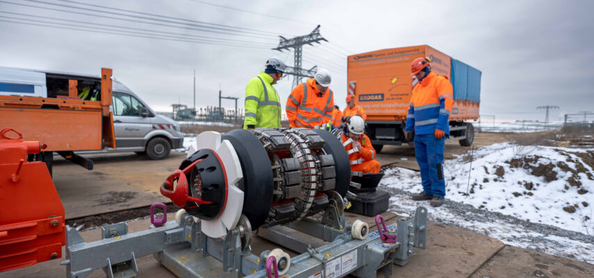 Arbeiter in Warnschutzkleidung prüfen ein großes Rohrbohrwerk auf einer verschneiten Baustelle vor orangefarbenem Firmen-Lkw und Stromleitungen im Hintergrund.