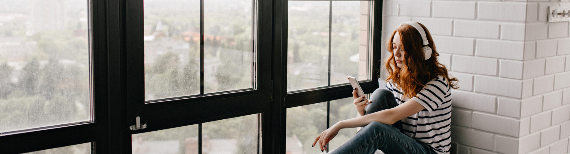 Frau mit roten Haaren und Kopfhörern sitzt im gestreiften Shirt auf einer Fensterbank und blickt auf ihr Smartphone.