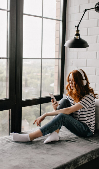 Frau mit roten Haaren und Kopfhörern sitzt im gestreiften Shirt auf einer Fensterbank und blickt auf ihr Smartphone.