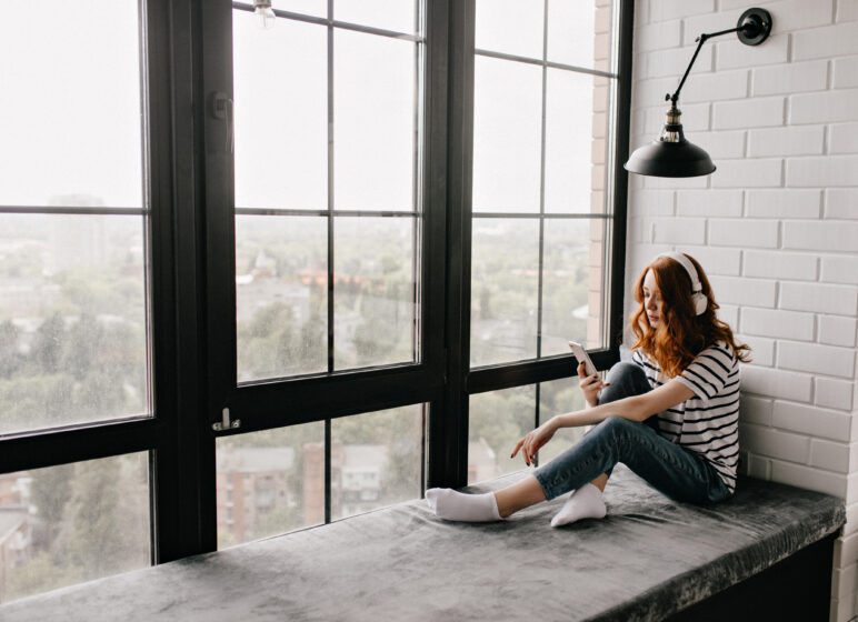 Frau mit roten Haaren und Kopfhörern sitzt im gestreiften Shirt auf einer Fensterbank und blickt auf ihr Smartphone.