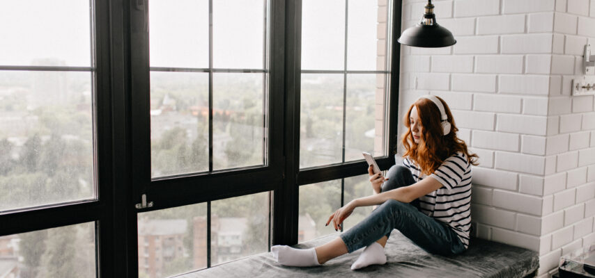 Frau mit roten Haaren und Kopfhörern sitzt im gestreiften Shirt auf einer Fensterbank und blickt auf ihr Smartphone.