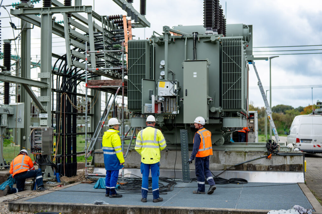 Mehrere Arbeiter in Warnwesten und Schutzhelmen arbeiten an einem großen Umspanntransformator mit Kabeln und Schaltschränken.