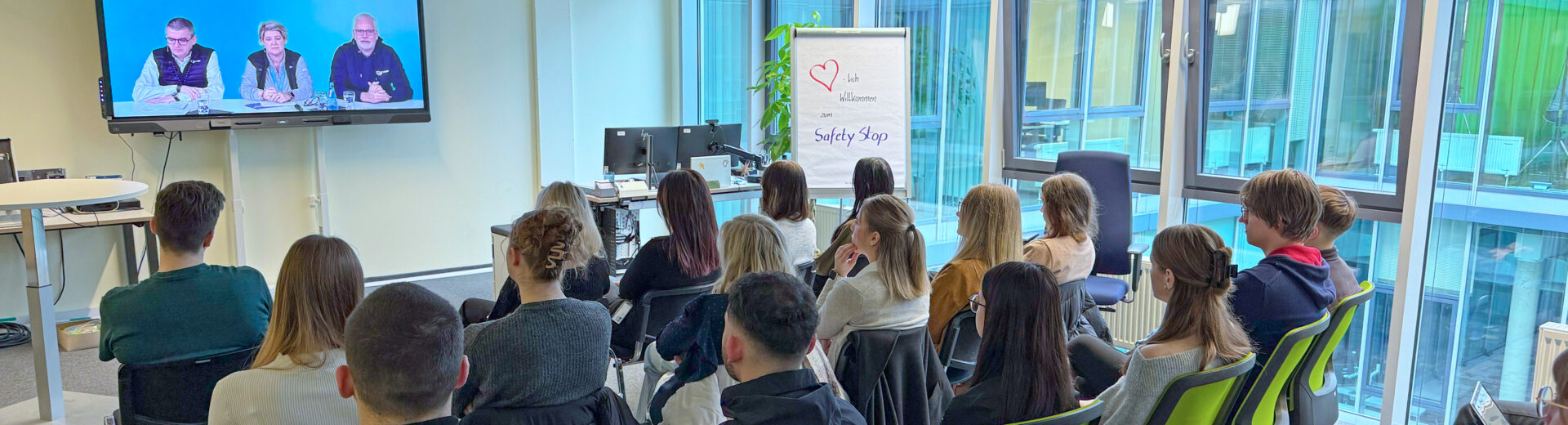 Teilnehmende sitzen auf grünen Stühlen in einem Büro und sehen eine Videopräsentation, Flipchart zeigt Safety Stop.