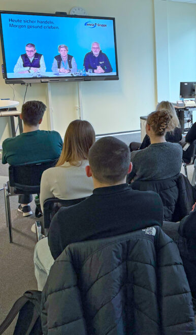Teilnehmende sitzen auf grünen Stühlen in einem Büro und sehen eine Videopräsentation, Flipchart zeigt Safety Stop.
