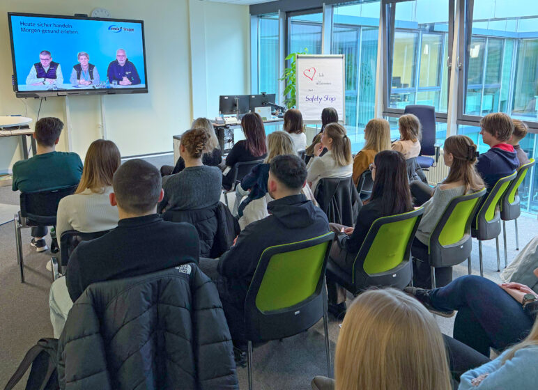 Teilnehmende sitzen auf grünen Stühlen in einem Büro und sehen eine Videopräsentation, Flipchart zeigt Safety Stop.