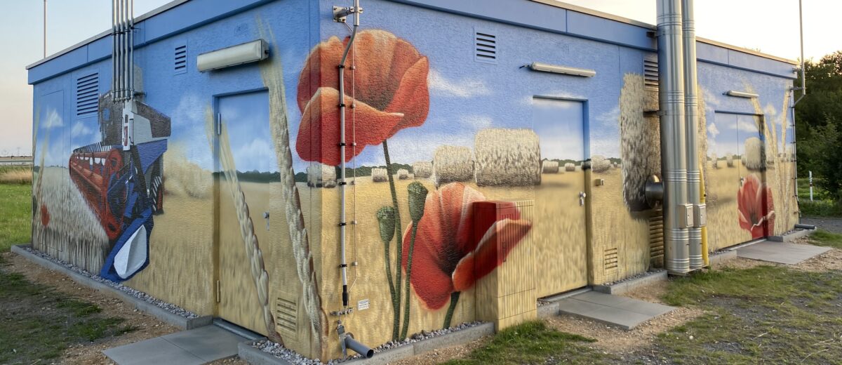 Versorgungsgebäude mit großflächigem Wandbild eines Weizenfelds mit Strohballen, roten Mohnblumen und Mähdrescher.