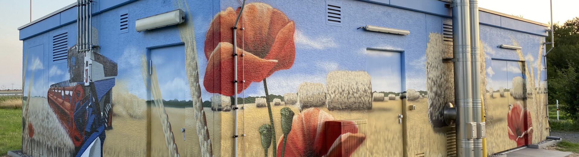 Versorgungsgebäude mit großflächigem Wandbild eines Weizenfelds mit Strohballen, roten Mohnblumen und Mähdrescher.