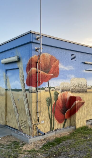 Versorgungsgebäude mit großflächigem Wandbild eines Weizenfelds mit Strohballen, roten Mohnblumen und Mähdrescher.