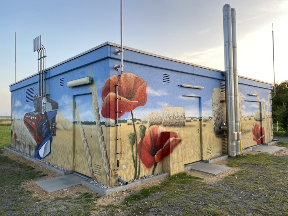 Versorgungsgebäude mit großflächigem Wandbild eines Weizenfelds mit Strohballen, roten Mohnblumen und Mähdrescher.