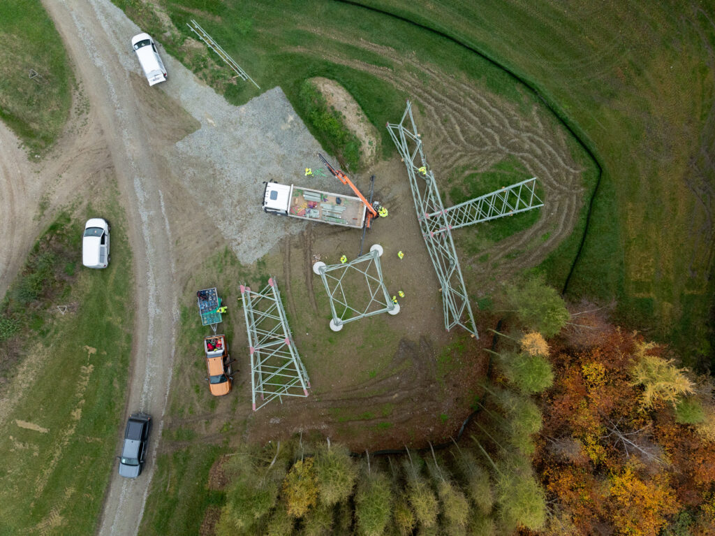Bauarbeiter und Fahrzeuge beim Zusammensetzen mehrerer Strommasten auf einer Wiesenschneise, aufgenommen aus der Vogelperspektive.