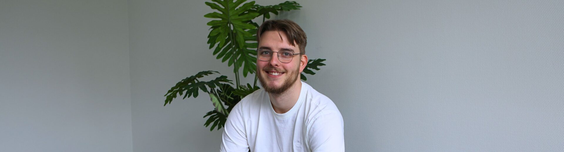 Junger Mann mit Brille und Bart sitzt lächelnd auf grauem Sofa, trägt weißes T‑Shirt, beige Hose und schwarze Uhr; Pflanze im Hintergrund.