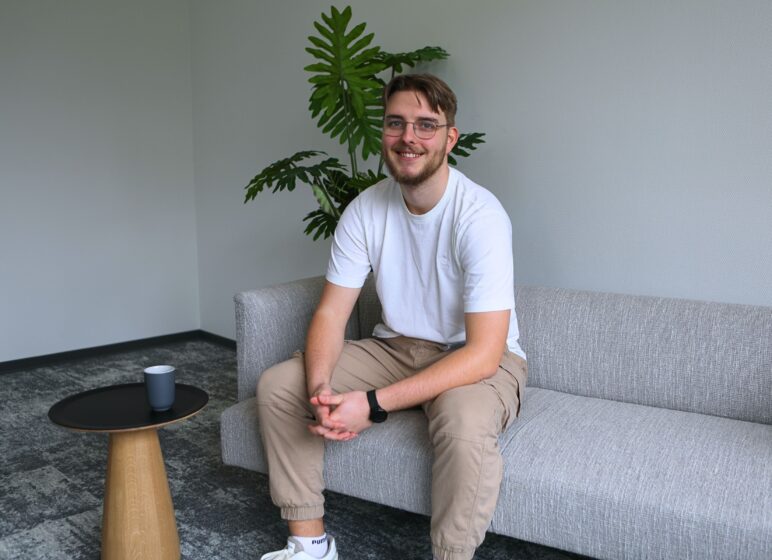 Junger Mann mit Brille und Bart sitzt lächelnd auf grauem Sofa, trägt weißes T‑Shirt, beige Hose und schwarze Uhr; Pflanze im Hintergrund.