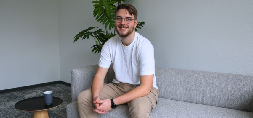 Junger Mann mit Brille und Bart sitzt lächelnd auf grauem Sofa, trägt weißes T‑Shirt, beige Hose und schwarze Uhr; Pflanze im Hintergrund.