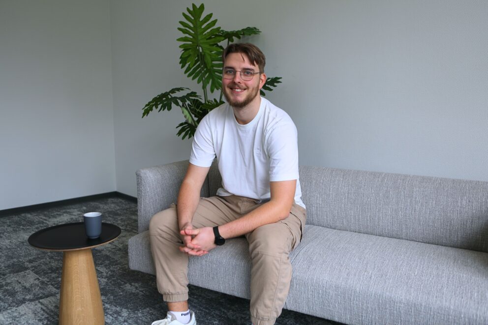 Junger Mann mit Brille und Bart sitzt lächelnd auf grauem Sofa, trägt weißes T‑Shirt, beige Hose und schwarze Uhr; Pflanze im Hintergrund.