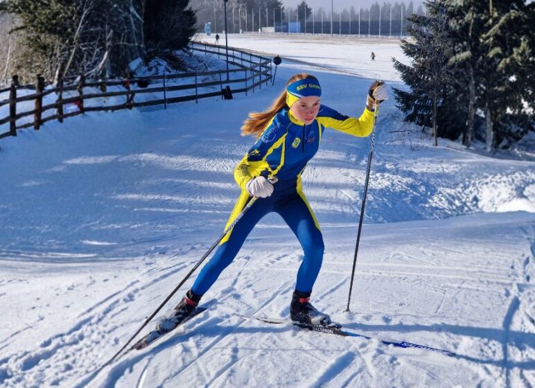 Skilangläuferin in blau-gelbem Rennanzug und Stirnband fährt mit Skiern und Stöcken über verschneiten Hang neben Holzzaun.