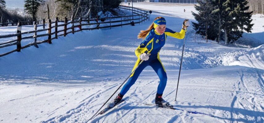 Skilangläuferin in blau-gelbem Rennanzug und Stirnband fährt mit Skiern und Stöcken über verschneiten Hang neben Holzzaun.