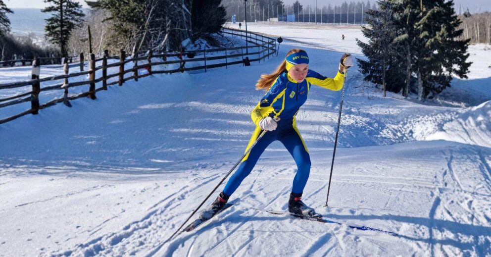 Skilangläuferin in blau-gelbem Rennanzug und Stirnband fährt mit Skiern und Stöcken über verschneiten Hang neben Holzzaun.