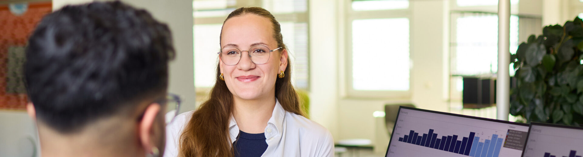 Lächelnde Beraterin mit Brille und weißer Bluse im Gespräch mit Mann in blauem Hoodie, Monitore zeigen Balkendiagramme.