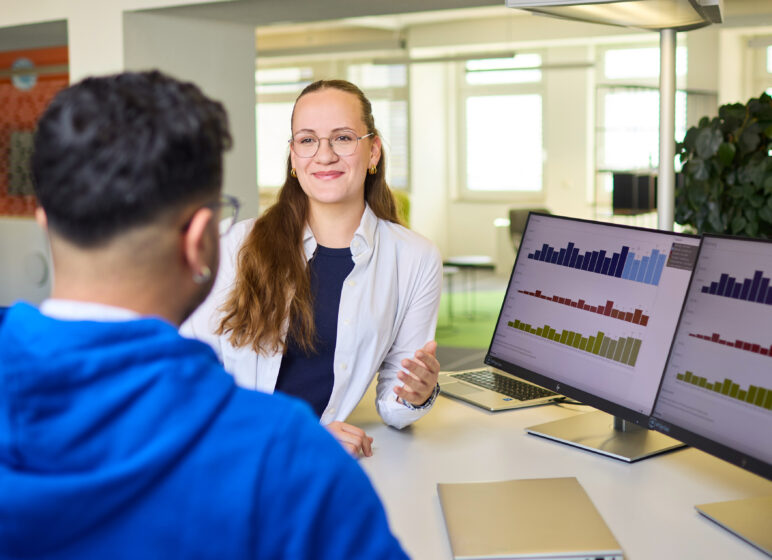 Lächelnde Beraterin mit Brille und weißer Bluse im Gespräch mit Mann in blauem Hoodie, Monitore zeigen Balkendiagramme.