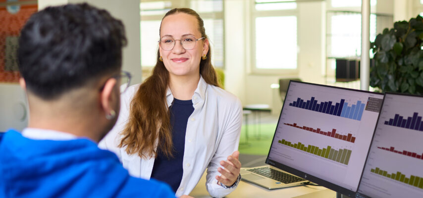 Lächelnde Beraterin mit Brille und weißer Bluse im Gespräch mit Mann in blauem Hoodie, Monitore zeigen Balkendiagramme.