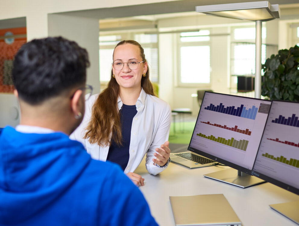 Lächelnde Beraterin mit Brille und weißer Bluse im Gespräch mit Mann in blauem Hoodie, Monitore zeigen Balkendiagramme.