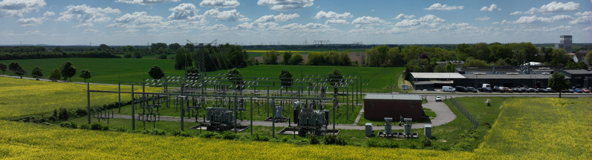 Weites Panorama mit gelbem Rapsfeld im Vordergrund, Umspannwerk in der Bildmitte und blauem Himmel mit weißen Wolken.