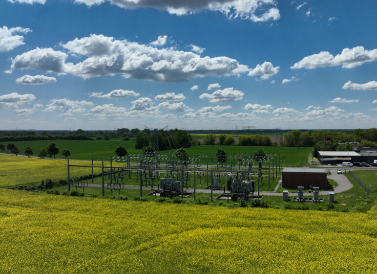 Weites Panorama mit gelbem Rapsfeld im Vordergrund, Umspannwerk in der Bildmitte und blauem Himmel mit weißen Wolken.