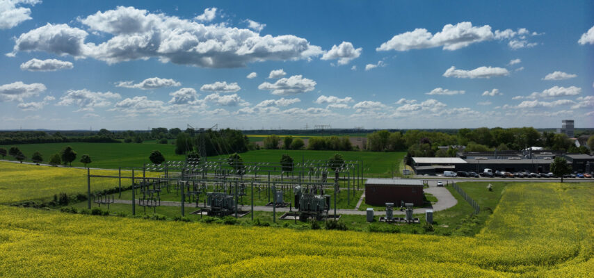 Weites Panorama mit gelbem Rapsfeld im Vordergrund, Umspannwerk in der Bildmitte und blauem Himmel mit weißen Wolken.