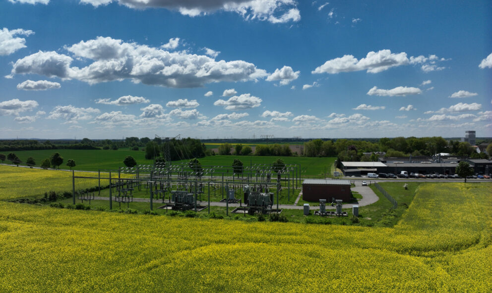 Weites Panorama mit gelbem Rapsfeld im Vordergrund, Umspannwerk in der Bildmitte und blauem Himmel mit weißen Wolken.