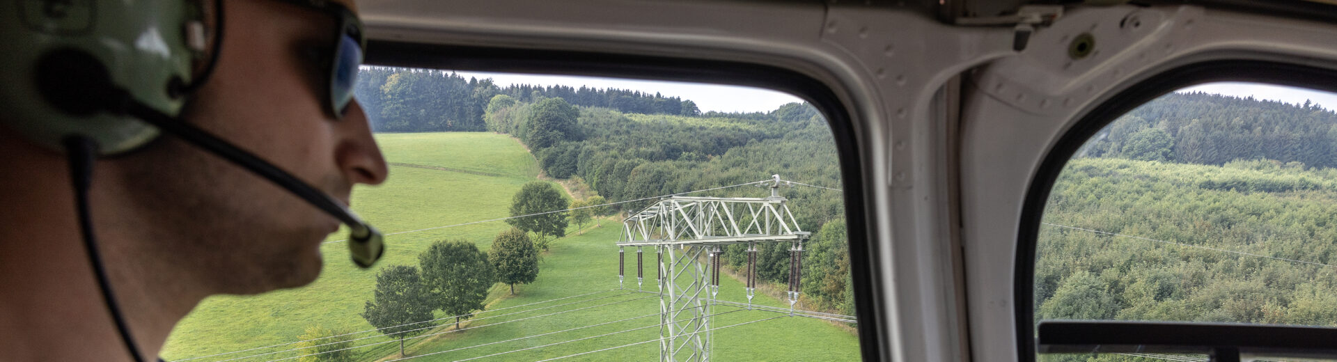 Pilot mit Headset blickt aus einem Helikopterfenster auf einen Strommast über landwirtschaftlichen Feldern und Hügeln, Aktenmappe im Schoß.
