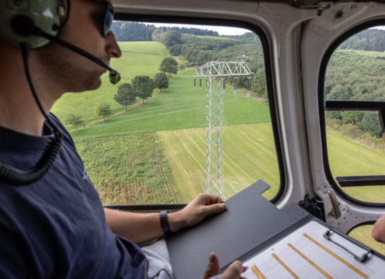Pilot mit Headset blickt aus einem Helikopterfenster auf einen Strommast über landwirtschaftlichen Feldern und Hügeln, Aktenmappe im Schoß.