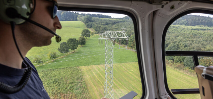 Pilot mit Headset blickt aus einem Helikopterfenster auf einen Strommast über landwirtschaftlichen Feldern und Hügeln, Aktenmappe im Schoß.
