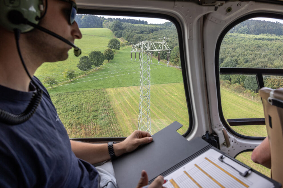 Pilot mit Headset blickt aus einem Helikopterfenster auf einen Strommast über landwirtschaftlichen Feldern und Hügeln, Aktenmappe im Schoß.