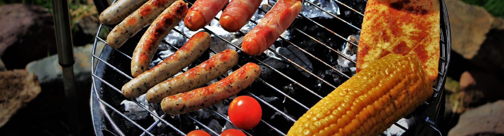 Grill über Kohlen mit verschiedenen Bratwürsten, Speckwürstchen, gegrilltem Maiskolben, Tomaten, Toastbrötchen und gebratenem Käse.