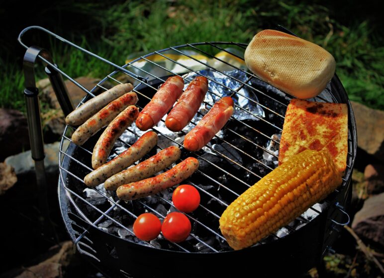 Grill über Kohlen mit verschiedenen Bratwürsten, Speckwürstchen, gegrilltem Maiskolben, Tomaten, Toastbrötchen und gebratenem Käse.