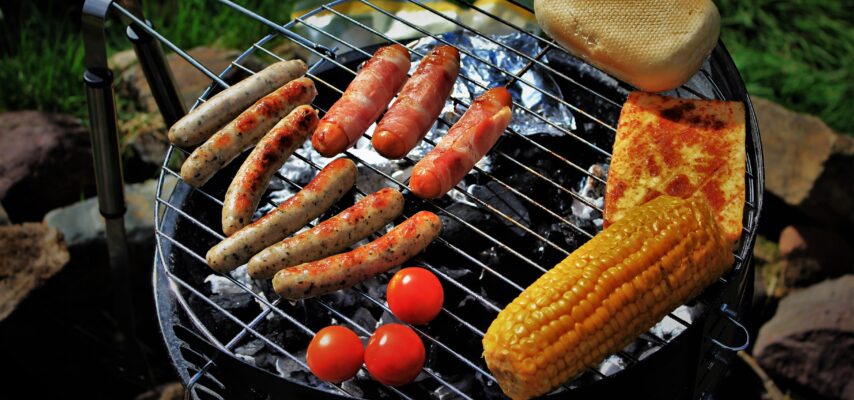 Grill über Kohlen mit verschiedenen Bratwürsten, Speckwürstchen, gegrilltem Maiskolben, Tomaten, Toastbrötchen und gebratenem Käse.