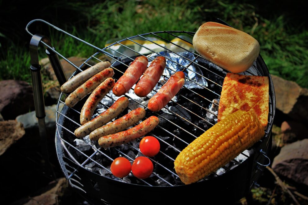 Grill über Kohlen mit verschiedenen Bratwürsten, Speckwürstchen, gegrilltem Maiskolben, Tomaten, Toastbrötchen und gebratenem Käse.