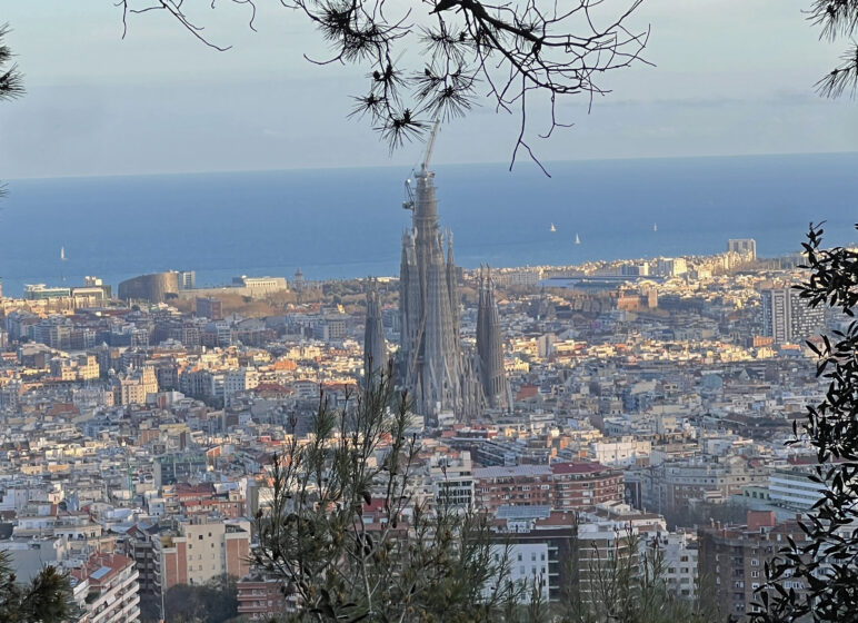 Sicht auf Barcelona mit der Sagrada Família im Vordergrund, blaues Meer im Hintergrund und Bäume als Rahmen.