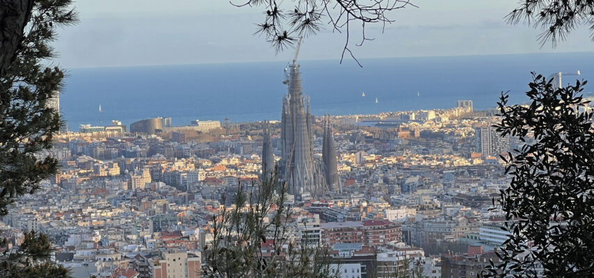 Sicht auf Barcelona mit der Sagrada Família im Vordergrund, blaues Meer im Hintergrund und Bäume als Rahmen.