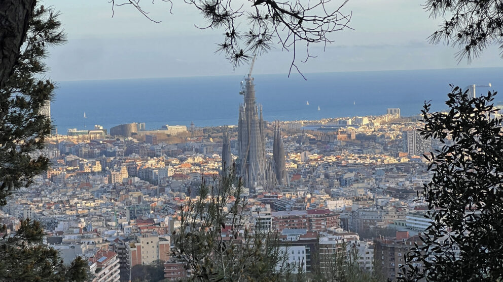 Sicht auf Barcelona mit der Sagrada Família im Vordergrund, blaues Meer im Hintergrund und Bäume als Rahmen.