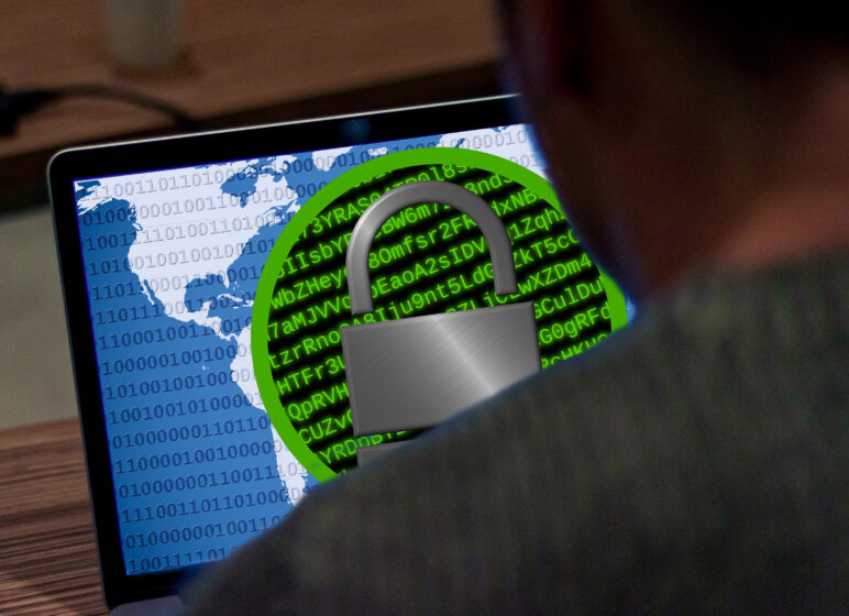 Laptopbildschirm mit Weltkarte, Binärcode und großem Vorhängeschloss-Symbol vor grünem Codehintergrund, Person im Vordergrund.